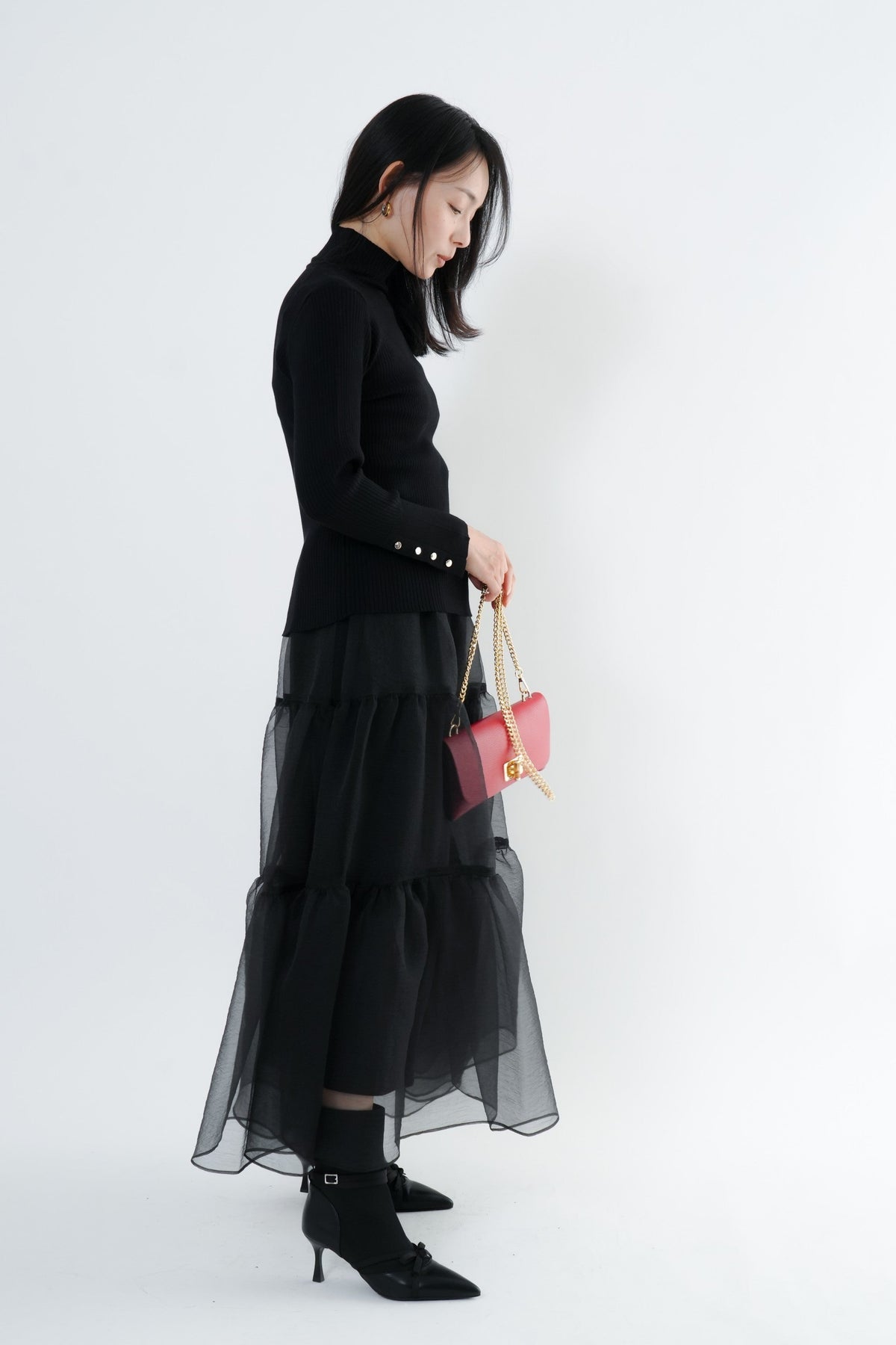 e-clothing-online storeのCLOCHEリブハイネックニットを着用した女性。袖口のゴールドボタンが特徴の黒いタートルネックニットに黒のチュールスカートを合わせた上品なスタイル。