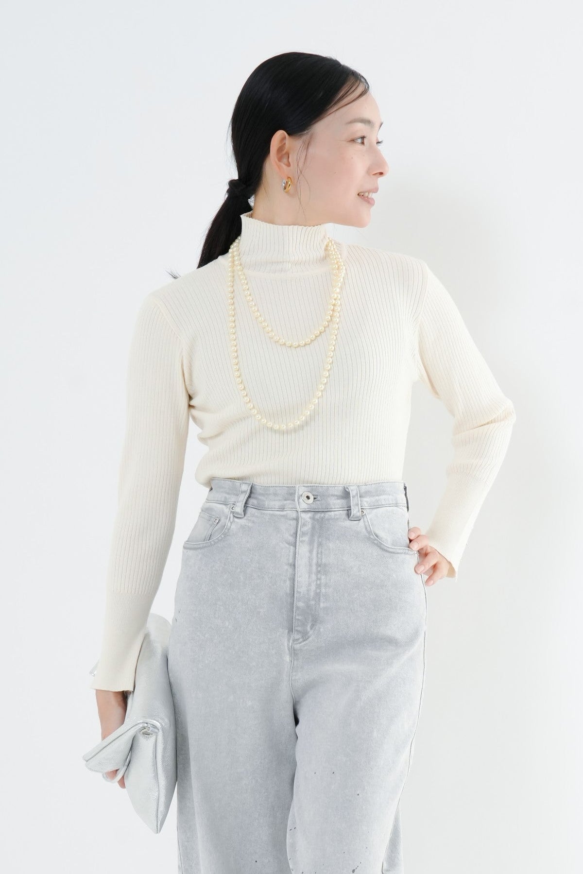 e-clothing-online storeのCLOCHEリブハイネックニットを着用した女性。袖口のゴールドボタンが特徴のホワイトのタートルネックニットに、ライトグレーのデニムを合わせたきれいめな秋冬スタイル。